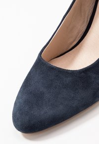 Tummansininen mokkanahkainen korkokengät, joissa on terävä kärki ja kaareva aukko. Suuri tekstuuri, minimalistinen muotoilu ja neutraali nahkasisus.