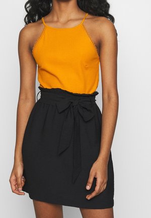 Femme portant un haut orange sans manches en maille côtelée avec bordure en dentelle et une jupe noire taille haute nouée à la taille avec un nœud de ceinture en tissu.