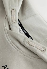 Beiger Kapuzenpullover mit Kordelzug, der durch Metallösen geführt wird, sichtbaren Nähten und einem schwarzen AllSaints Markenlabel innen am Kragen.