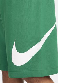 Zelené látkové šortky s velkým bílým logem Nike swoosh na spodní straně, nošené na noze člověka.