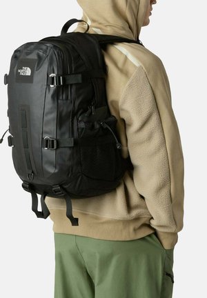 Zwarte rugzak met verstelbare riemen, meerdere vakken en een zijvak van gaas. Heeft een gladde, glanzende afwerking en een logodetail aan de voorkant.