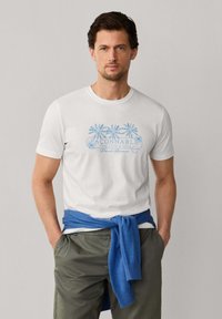 Mann trägt weißes T-Shirt mit blauem Palmenprint und Schriftzug, olivgrüne Hose und blauen Pullover um die Taille gebunden, steht mit Händen in den Taschen.