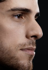 Profil d'un visage masculin avec un teint lisse, un nez proéminent et une légère barbe. Yeux marron et cheveux foncés sur fond noir.
