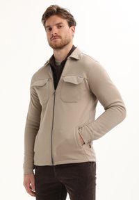 Hombre vistiendo una chaqueta beige con cremallera y bolsillos en el pecho sobre una camisa y pantalones marrón oscuro, posando con una mano en el bolsillo frente a un fondo blanco.