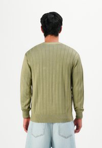 Homem de costas, vestindo um suéter canelado verde claro e calções de ganga azul-claro, contra um fundo branco.