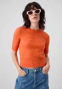 Krátky rukáv, oranžový pletený sveter s rebrovaným výstrihom a lemom. V kombinácii s modrou džínsovou sukňou, model pózuje s rukami vo vreckách.