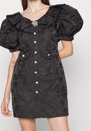Mini-robe noire texturée avec manches bouffantes, broche florale et boutons bijoux sur le devant, avec deux petites poches avant.