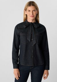 Donkerdenim overhemd met lange mouwen, knoopsluiting aan de voorkant, twee borstzakken en een strikceintuur detail. Contrasterende oranje stiksels accentueren de randen.