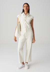 Combinaison en jean blanche à manches courtes, fermeture à boutons, poche frontale et coupe ample, associée à des baskets blanches et un sac en tissu.