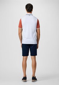 Witte polo met rode mouwen, marineblauwe shorts en zwarte schoenen. Heeft een hoge kraag en een eenvoudig logo op de nek. Gladde textuur.