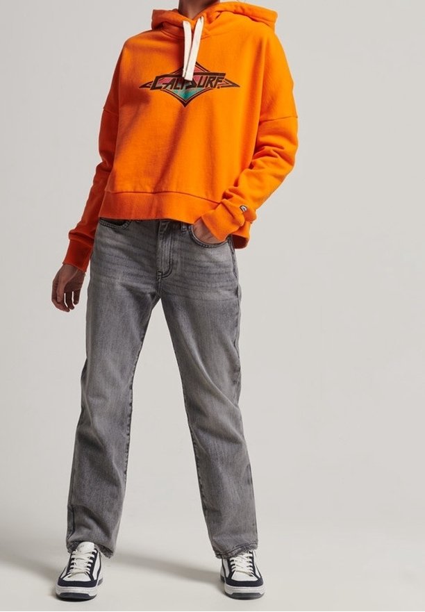 Personne debout, les mains dans les poches, portant un sweat à capuche orange vif avec logo, un jean gris et des baskets noir et blanc sur fond uni.