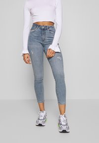 Jeans de tiro alto azul claro con detalles desgastados, corte ajustado y longitud por encima del tobillo, combinados con una camiseta blanca de manga larga y zapatillas grises.