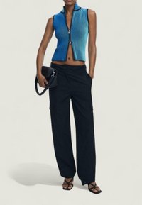 Vrouw in een mouwloze blauwe zip-up crop top, zwarte wijde broek, zwarte hakken met banden, en met een zwarte handtas met een kettingband.