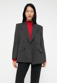 Frau, die ein dunkelgraues, gestreiftes, doppelt geknöpftes Blazer über einem roten Rollkragenpullover und schwarzen Hosen trägt, vor einem weißen Hintergrund steht.