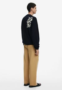 Marineblå strikket sweater med struktureret stof og cremefarvet grafisk tekst på ryggen, parret med beige løstsiddende bukser og sorte sandaler.