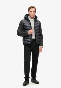 Giacca imbottita nera da uomo con cappuccio e maniche in maglia grigio scuro testurizzato, abbinata a un maglione grigio chiaro con zip a quarto e pantaloni neri.