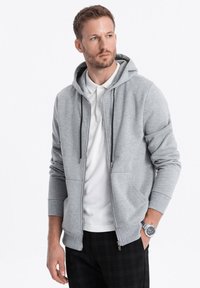 Sweat à capuche zippé gris clair en mélange de coton, avec deux poches avant, des cordons de serrage et des poignets côtelés. Porté par-dessus un polo blanc et un pantalon à carreaux.
