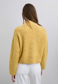 Pull en tricot texturé jaune avec un col montant et un ourlet côtelé. Coupe carrée, manches longues, vu de dos, associé à un pantalon blanc.