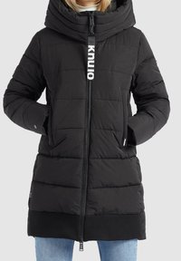 Mujer vestida con una chaqueta acolchada negra con un alto cuello, capucha oversized y un cierre largo con la etiqueta "knuio", con las manos en los bolsillos.