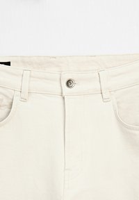 Pantaloni in denim di cotone color crema con chiusura a bottone e zip, dotati di cinque tasche, passanti per cintura e una texture liscia.