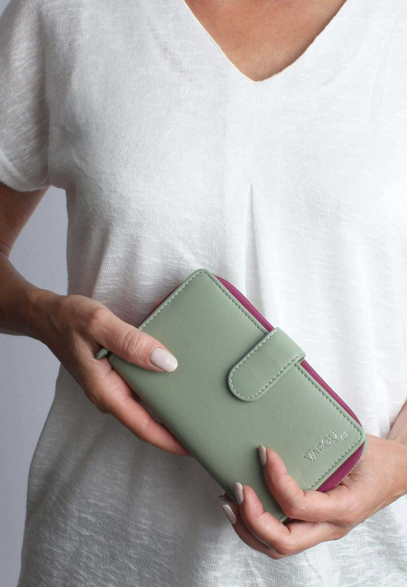 Green leather wallet with a pink interior, featuring a flap closure and stitching details. Held in hands against a light fabric background.