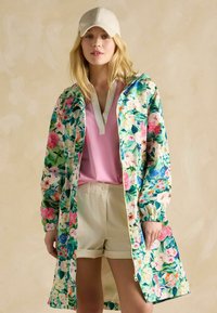 Lichte regenjas met bloemenpatroon in groen, roze en blauw, met een rits en elastische manchetten, gecombineerd met een roze top en beige shorts.