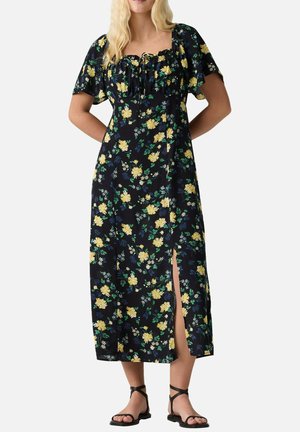 Frau trägt ein schwarzes, florales Midikleid mit gelben Blumen, kurzen Ärmeln, einem vorderen Schlitz und schwarzen flachen Riemchensandalen.