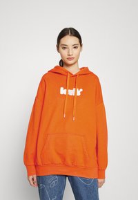 Joven vestida con una sudadera naranja brillante con un logo blanco y jeans azules que presentan dibujos de líneas de caras abstractas, de pie contra un fondo liso.