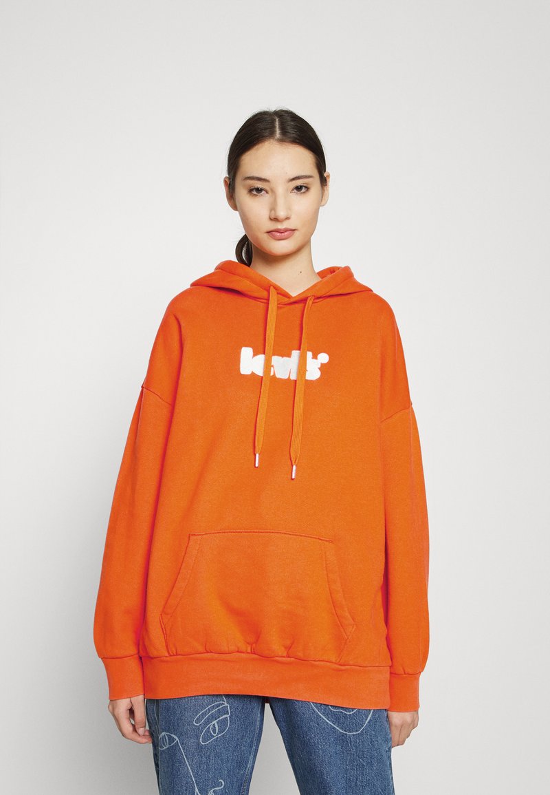 Joven vestida con una sudadera naranja brillante con un logo blanco y jeans azules que presentan dibujos de líneas de caras abstractas, de pie contra un fondo liso.