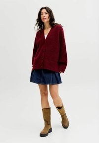 Burgunderfarbener Oversized-Cardigan mit V-Ausschnitt und Knopfverschluss, kombiniert mit einem Jeansrock und braunen kniehohen Stiefeln. Weicher Stoff mit lockerer Passform.