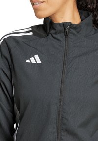 Schwarze Sportjacke aus leichtem Material. Verfügt über einen hohen Kragen, einen Reißverschluss in voller Länge und weiße Streifen auf den Schultern. Logo auf der Brust.