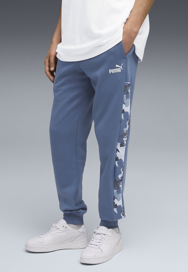 Blå sweatpants med et tekstureret camouflage sidepanel, hvidt logo og elastiske manchetter. Kombineret med hvide sneakers.