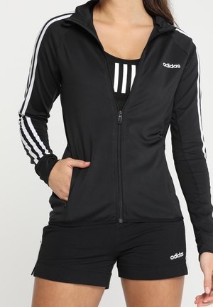 Femme portant une veste zippée noire Adidas avec des bandes blanches sur les manches, un soutien-gorge de sport noir et un short noir avec le logo Adidas.