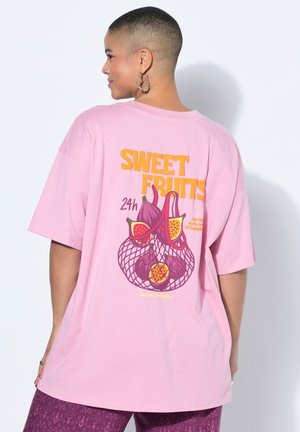 Osoba o krótkich włosach, ubrana w dużą różową koszulkę z napisem "Sweet Fruits" i grafiką owoców na plecach, w połączeniu z fioletowymi wzorzystymi spodniami.