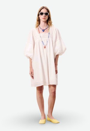 Femme portant une robe blanche à manches bouffantes, des colliers colorés en perles, des lunettes de soleil blanches et des chaussures plates jaunes, debout sur un fond uni.