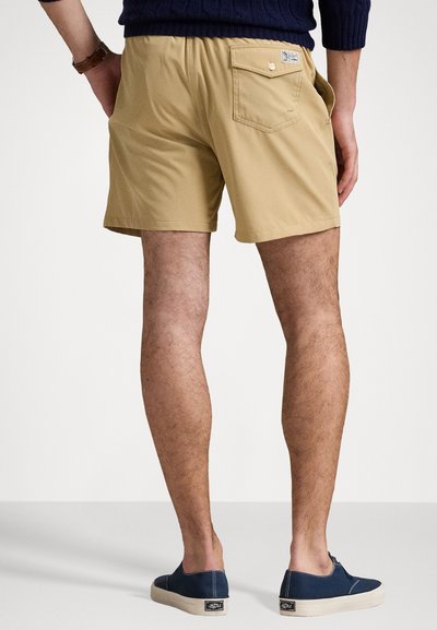 Bēšu šorti ar elastīgu jostu, aizmugures kabatu detaļu un gludu tekstūru, kopā ar tumši ziliem slip-on apaviem ar bēšu zoli.
