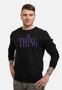 Giovane uomo con tatuaggi che indossa un felpa nera con la scritta viola "THING" e un'impronta di mano, in piedi con le mani sui fianchi su uno sfondo bianco.