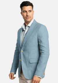Blazer de lino azul claro con un corte ajustado, dos botones y un bolsillo en el pecho. Se lleva sobre una camisa blanca estampada con cuello azul.