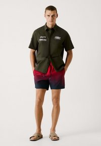 Tmavo zelená košeľa s krátkym rukávom a gombíkovým zapínaním, skombinovaná s plavkami v červeno-modrom prechode, ktoré obsahujú potlač textu. Outfit dopĺňajú béžové sandále.