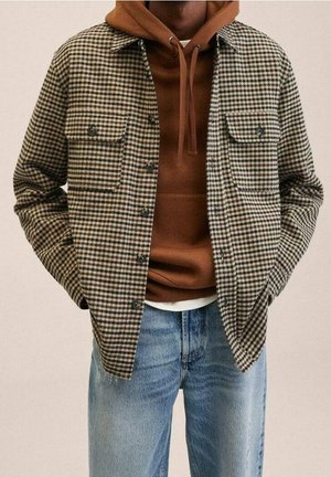 Mężczyzna ubrany w brązową bluzę z kapturem i sznurkami, brązowo-białą marynarkę w jodełkę zapinaną na guziki oraz jasno niebieskie dżinsy, ręce w kieszeniach marynarki.