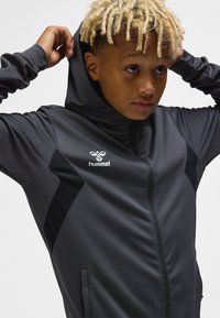 Junge Person mit blonden Dreadlocks zieht einen dunkelgrauen hummel Zip-up-Hoodie mit Kapuze vor einem schlichten hellen Hintergrund an.