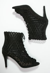 Calzado negro con un alto tacón de aguja, que presenta un patrón hexagonal cortado y un detalle de cordones en la parte delantera, fabricado en un material similar al ante.