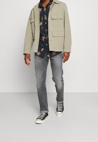 Chaqueta gris claro con múltiples bolsillos frontales, camisa navy con flores, jeans grises y zapatillas negras con suelas blancas. Conjunto casual.