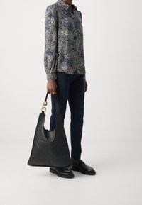 Sac en cuir noir avec une texture lisse et un dessus arrondi, doté de quincaillerie dorée et d'une large bandoulière, porté avec un pantalon sombre et une chemise à motifs.