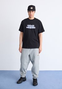 Ung mand står, iført en sort grafisk t-shirt, sort kasket, grå sweatpants og sorte træningssko på blå gulv mod en hvid væg.