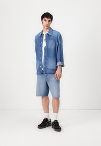 Lyseblå denimjakke med knapper, to frontlommer, hvit t-skjorte under, lyseblå denimshorts, svarte joggesko, og hvite sokker.