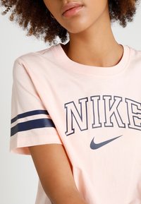 Světle růžové bavlněné tričko s námořnickým modrým logem "NIKE" a proužky na rukávech. Kulatý výstřih a volný střih. Měkká textura.