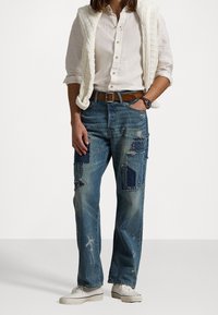 Světlé modré džínové kalhoty s nášivkami, odřenými částmi a volným střihem. Nošeny s bílou košilí na knoflíky a béžovou pletenou vestou.