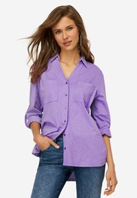 Camicia a maniche lunghe viola con colletto, due tasche anteriori e maniche arrotolate, realizzata in tessuto leggero; abbinata a jeans blu.