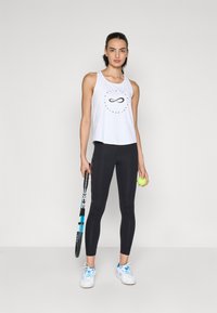 Top corto bianco senza maniche con un logo infinito nero, abbinato a leggings neri, scarpe da ginnastica bianche, mentre tiene una racchetta da tennis e una palla.